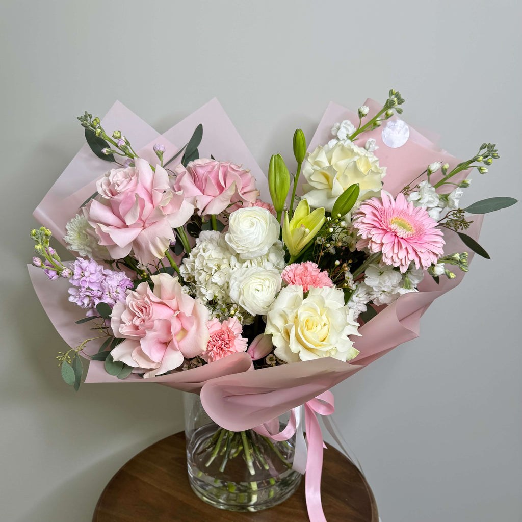 Charming bouquet of pink roses, white lilies, and ranunculus, wrapped in elegant pastel pink paper.