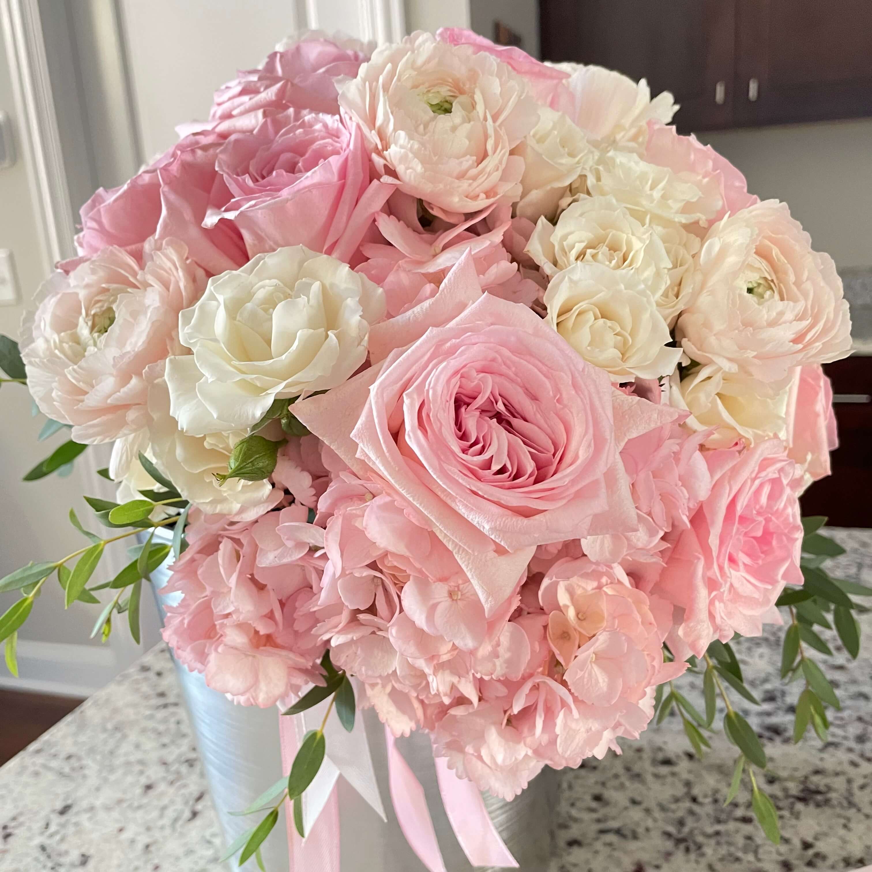 A pastel floral arrangement with pink roses, white carnations, and hydrangeas, delicately placed in a round box.