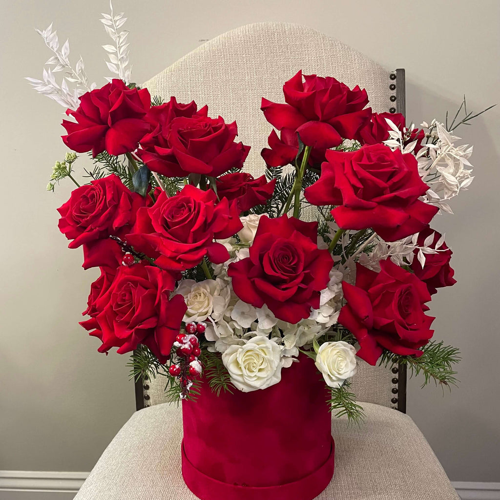 A luxurious floral arrangement featuring deep red roses, white hydrangeas, and white spray roses, elegantly placed in a red velvet hat box with festive greenery and decorative white branches.